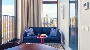 Un salon moderne avec un canapé bleu, une table ronde sur laquelle se trouve un bol de pop-corn et un verre, et une vue sur la ville grâce à de grandes fenêtres.