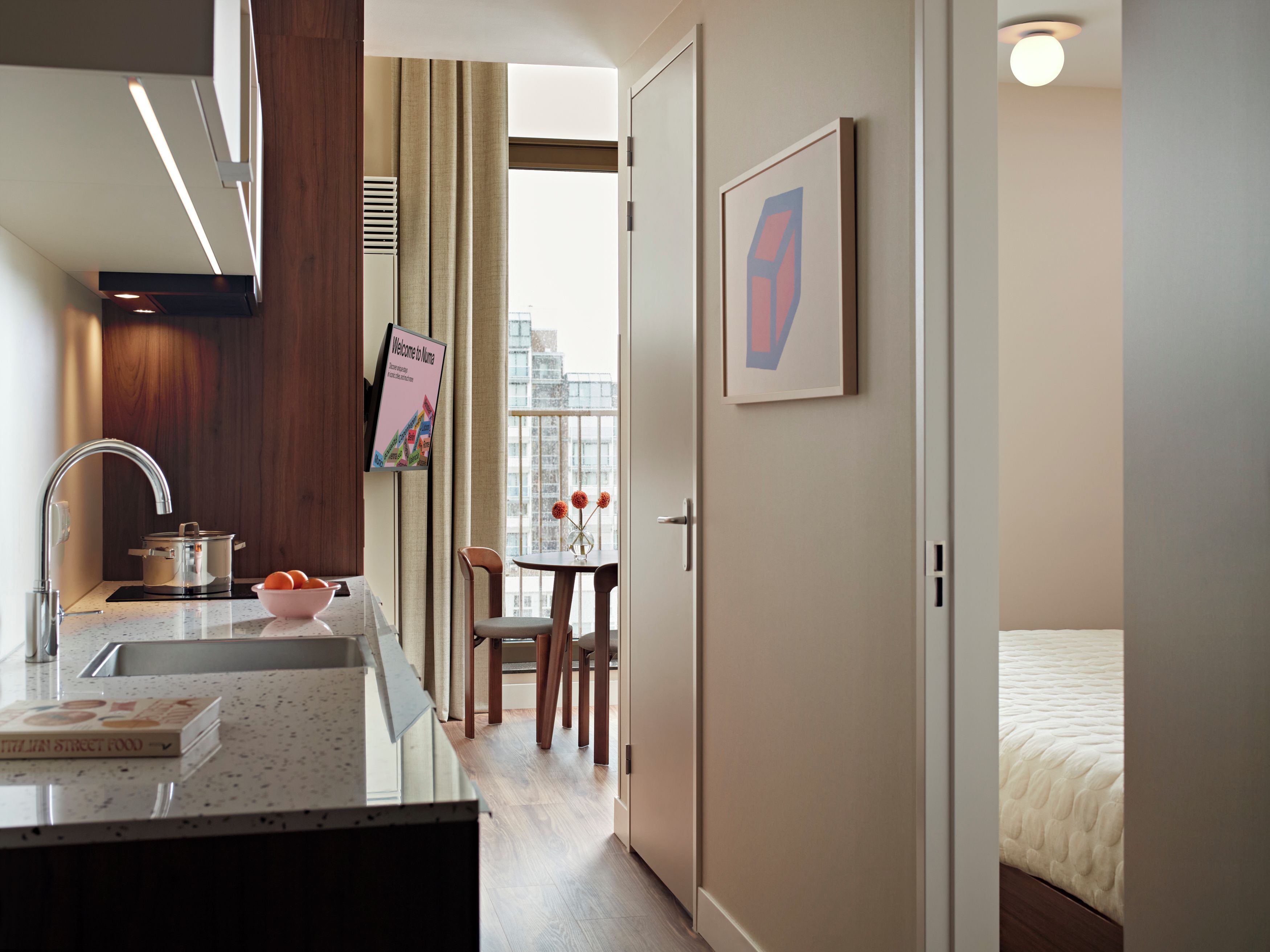 Ein modernes Hotelzimmer mit Küchenbereich, einem Esstisch mit Stühlen, einem Bett und einem gerahmten Bild an der Wand.