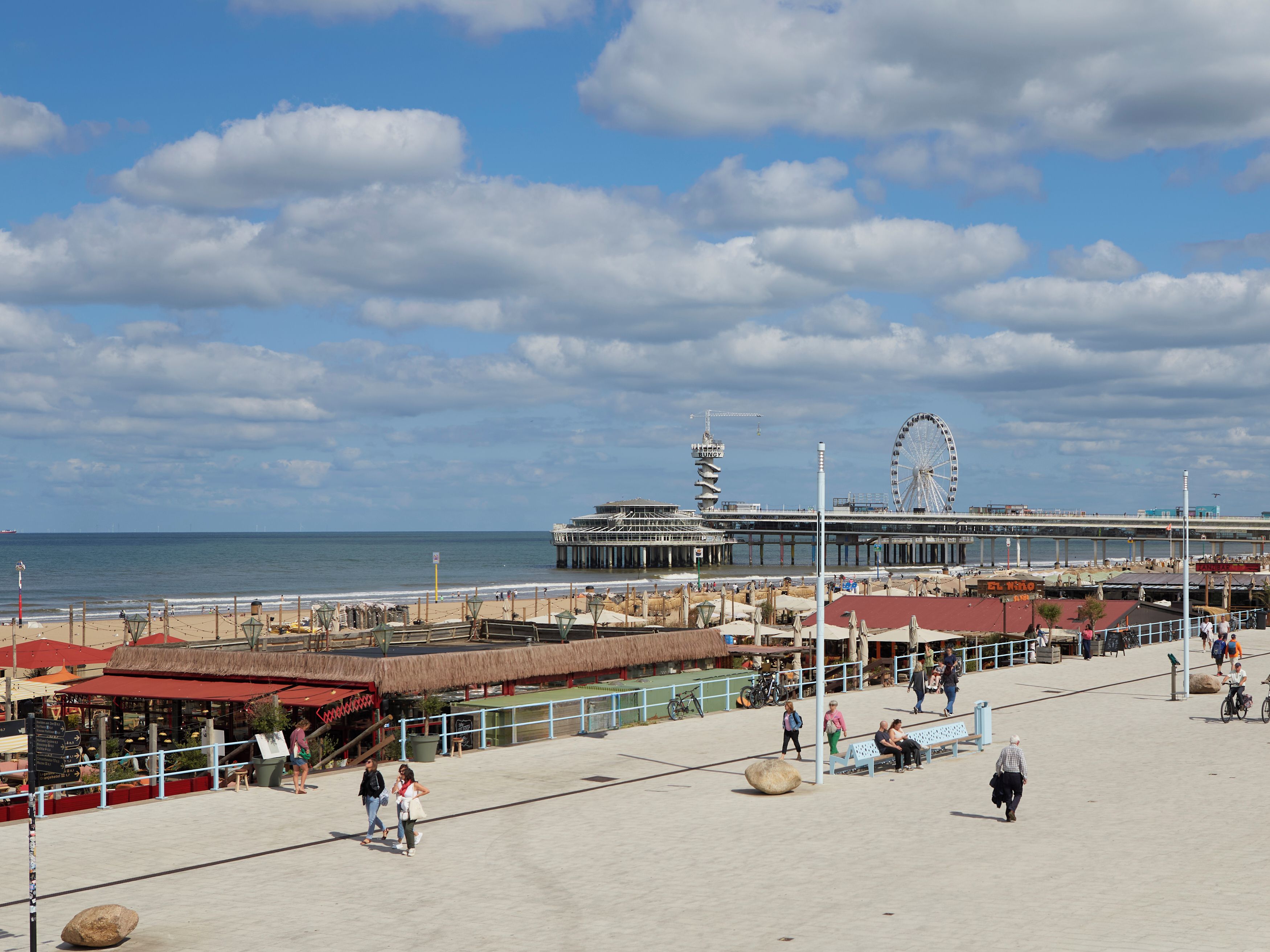 Un paseo marítimo frente a la playa con un muelle, noria y restaurante a pie de playa.