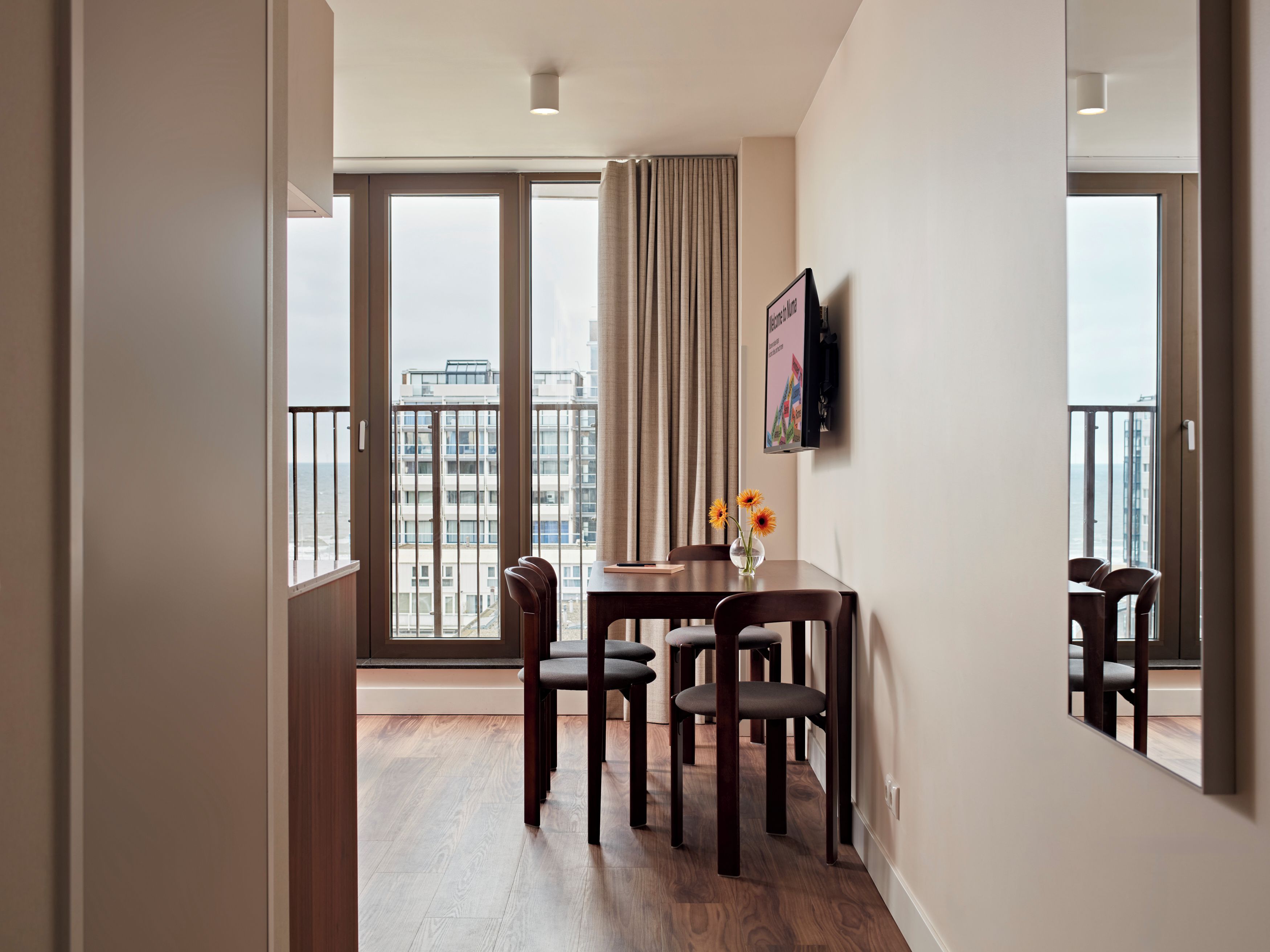 A modern hotel room with a balcony, a dining table with chairs, and a view of the cityscape.