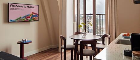 A modern hotel room with a dining area, a sofa, a small table with a vase, and a view of the city through large windows.