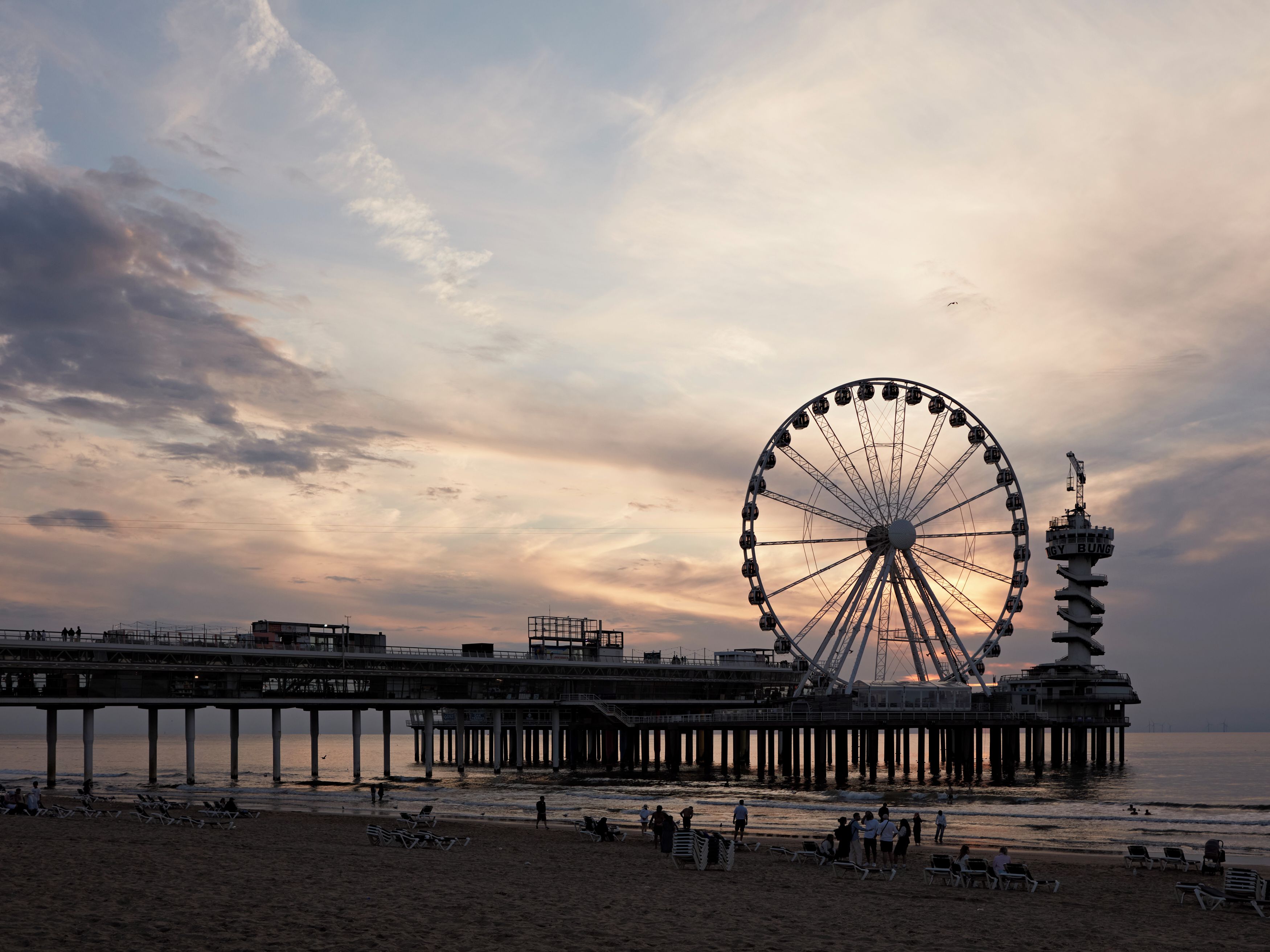 A pier with a large Ferris wheel, beach chairs, and people.