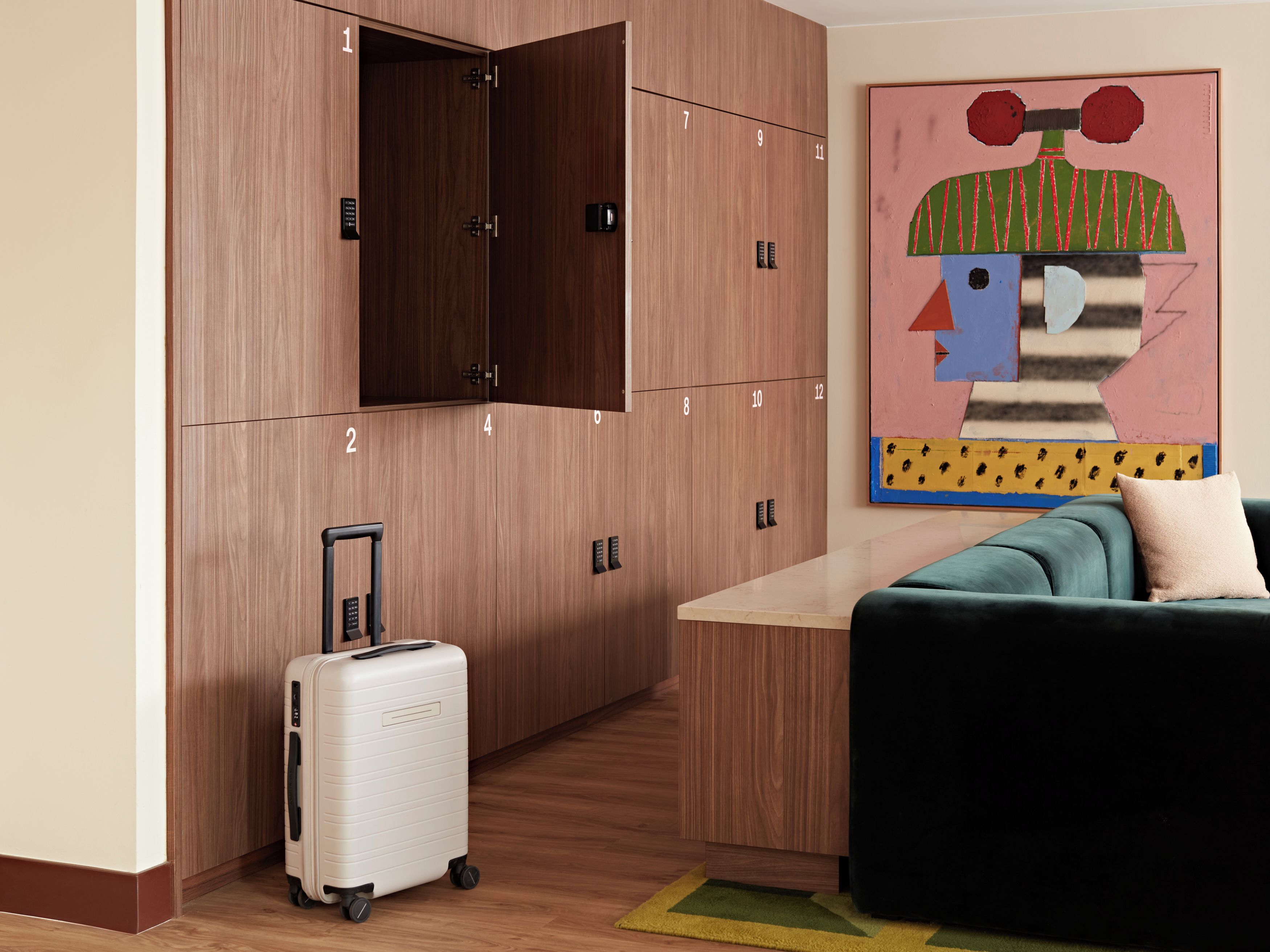 A hotel room with a wooden wardrobe, a single bed, a small table, a colorful abstract painting, and a suitcase.