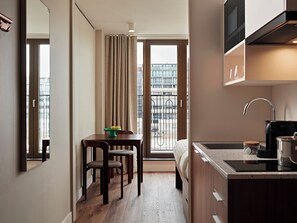 A compact hotel room with a kitchenette, a dining area with a table and chairs, a bed, and a large window with a view of the cityscape.
