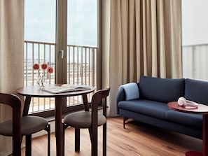 A modern living room with a blue sofa, a wooden dining table with chairs, and a view of a cityscape through large windows. - Numa The Hague Scheveningen (The Hague)