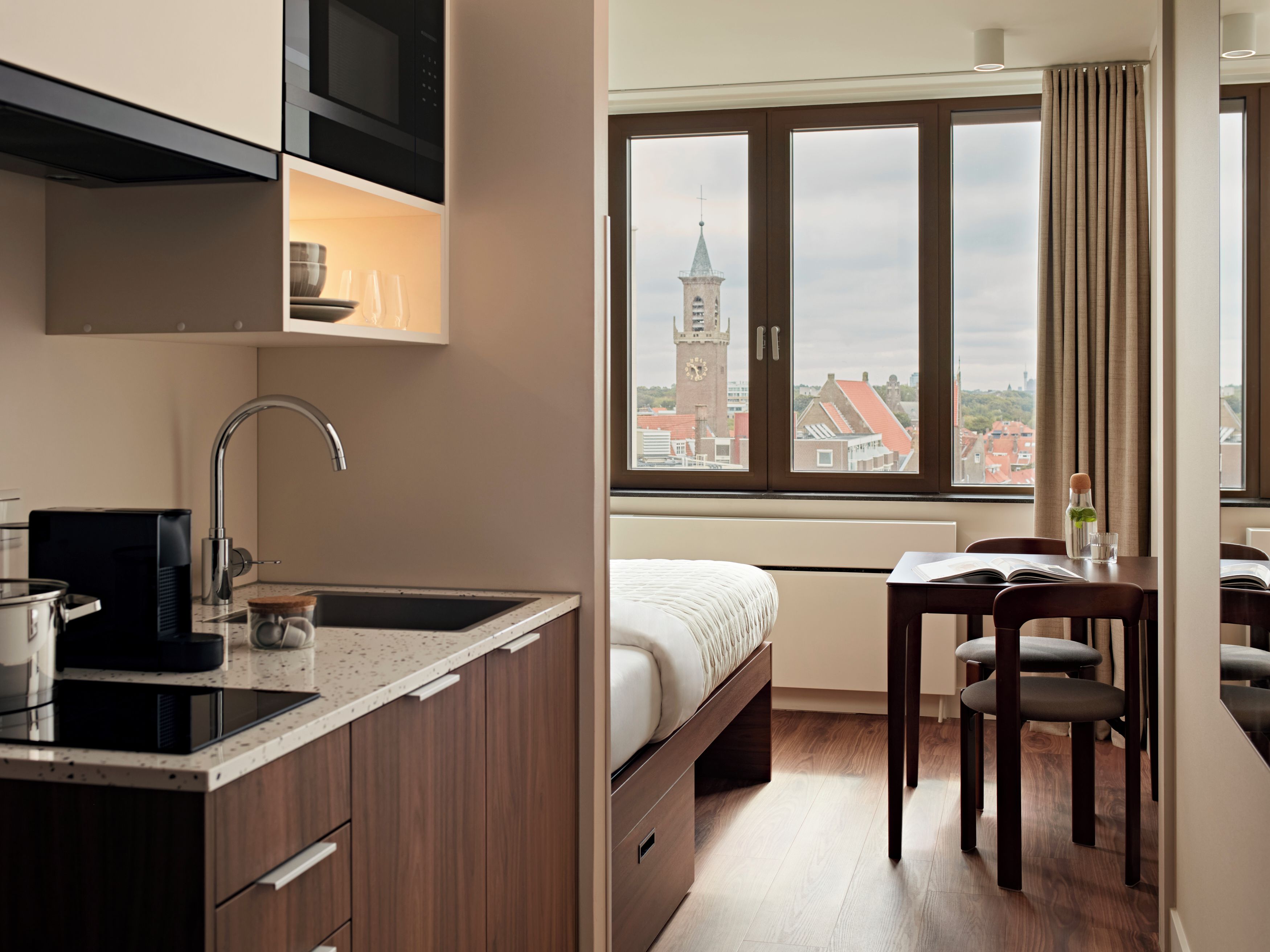 A compact hotel room with a kitchenette, a bed, a dining area, and a view of a clock tower through a window.