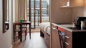 Habitación de hotel moderna con cama, zona de comedor, kitchenette y balcón con vistas a la ciudad.