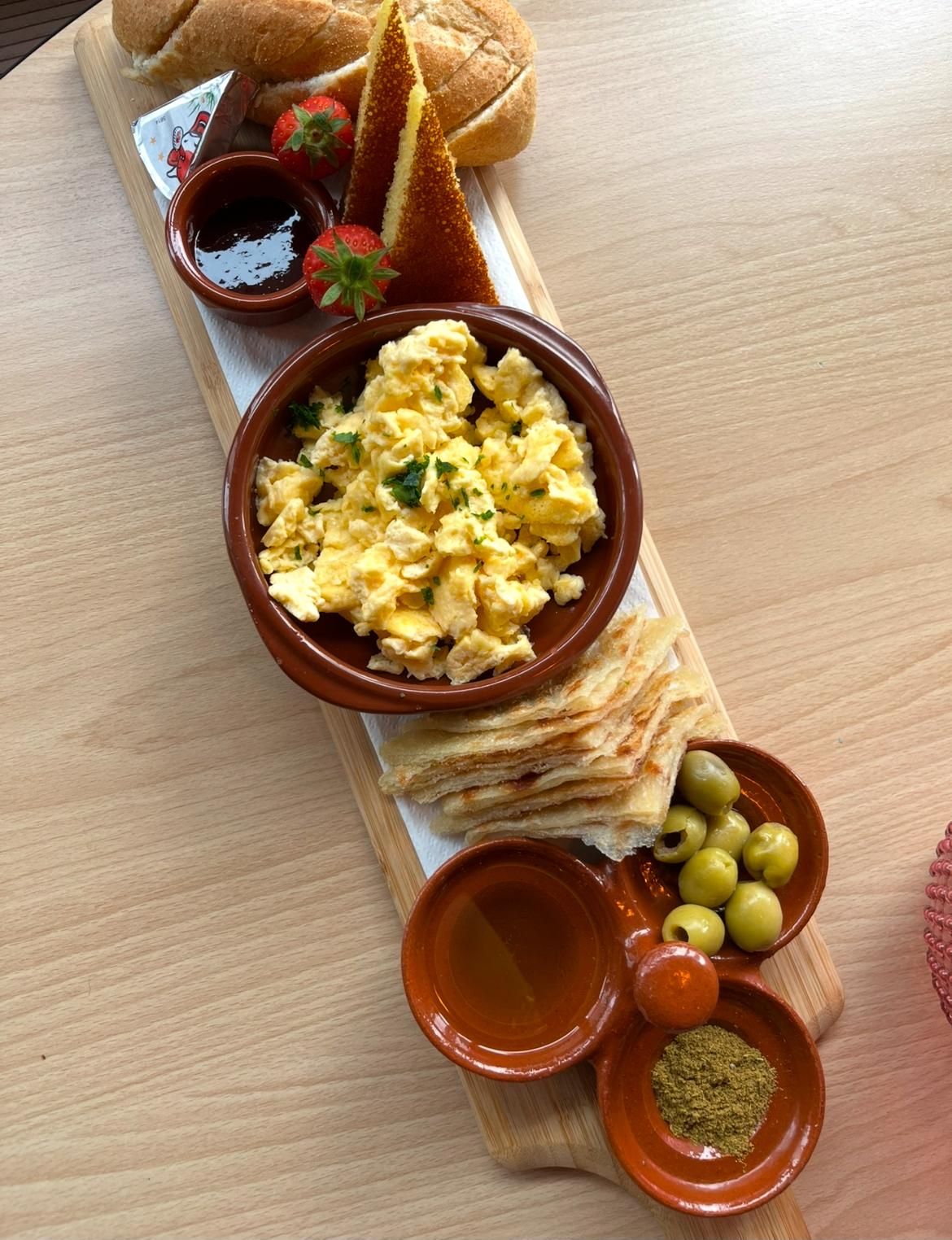 A wooden serving board with scrambled eggs, bread, olives, and condiments.