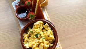 A wooden serving board with scrambled eggs, bread, olives, and condiments. - Numa The Hague Scheveningen (The Hague)
