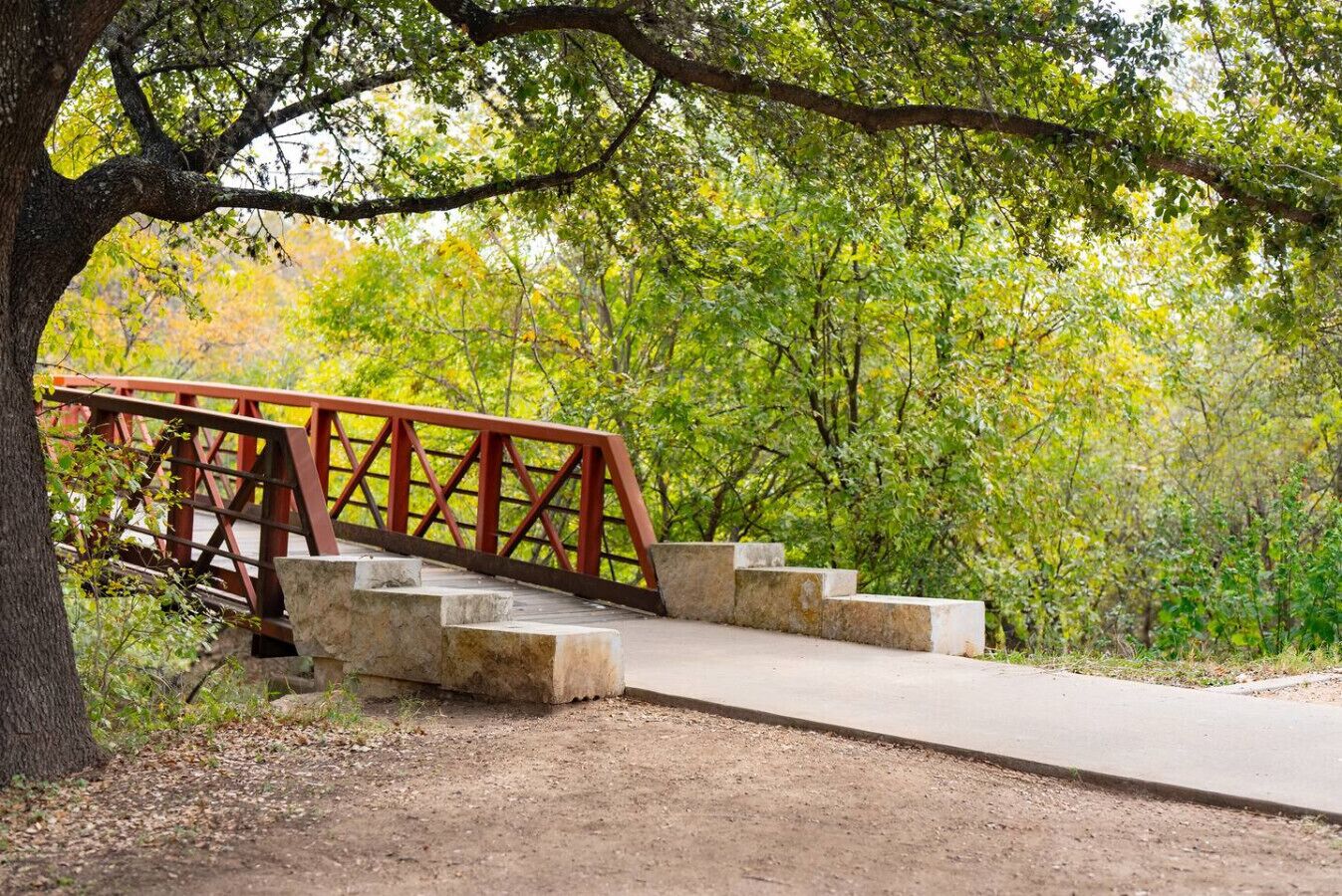 Eine Holzbrücke mit Steinstufen, die zu einem Betonweg in einem bewaldeten Gebiet führen.