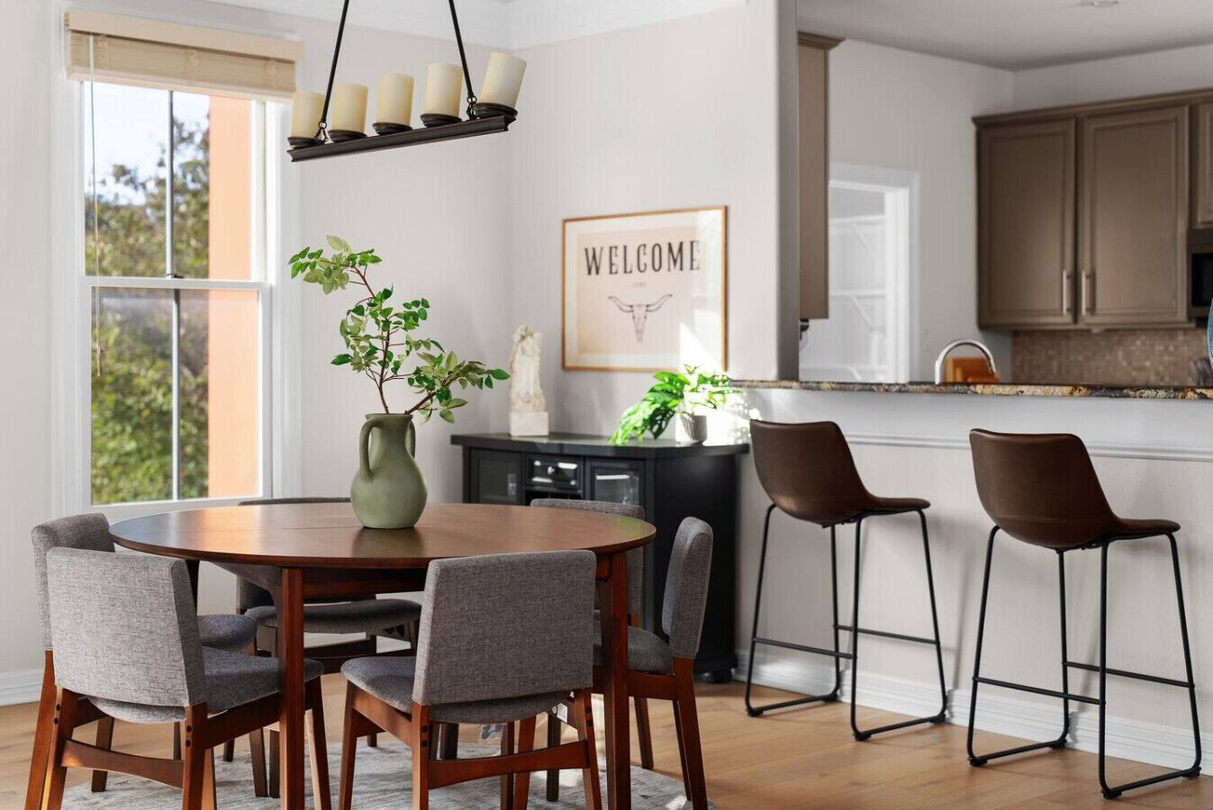 A dining area with a round table, four chairs, a vase with plants, a 'WELCOME' sign, and a kitchen in the background.