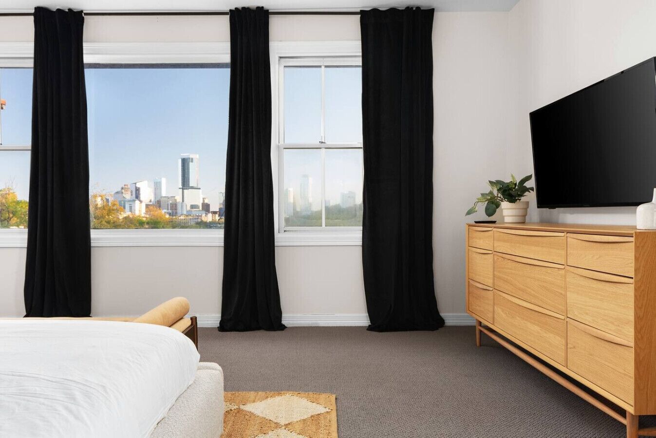 Ein modernes Hotelzimmer mit einem großen Bett, einer Holzkommode, einem Flachbildfernseher und Blick auf eine Stadtlandschaft durch raumhohe Fenster.