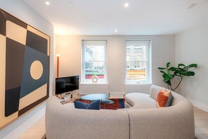 A modern living room with a sectional sofa, a flat-screen TV, two windows with city views, and a large abstract painting. - Apartment-sleeps4-parking-100walkingscore (London)