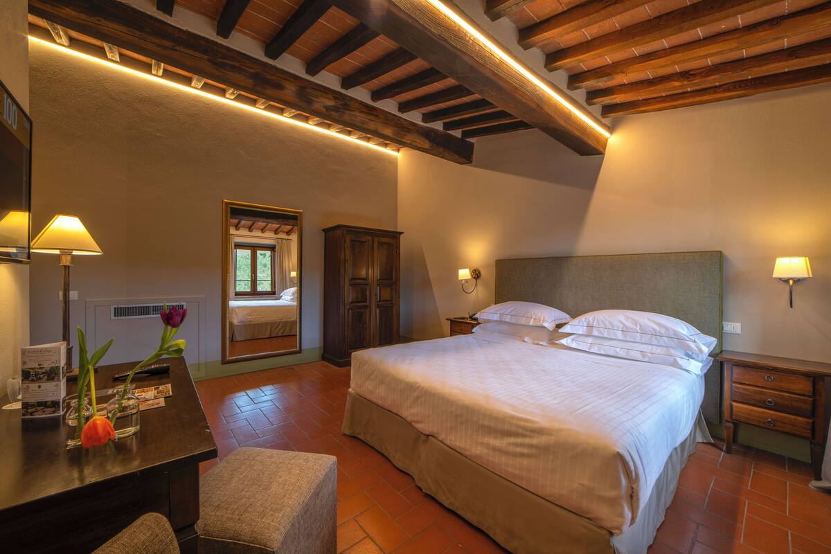 Une chambre à coucher avec un grand lit, des poutres apparentes en bois au plafond, une table de chevet et un bureau avec une lampe et des fleurs.