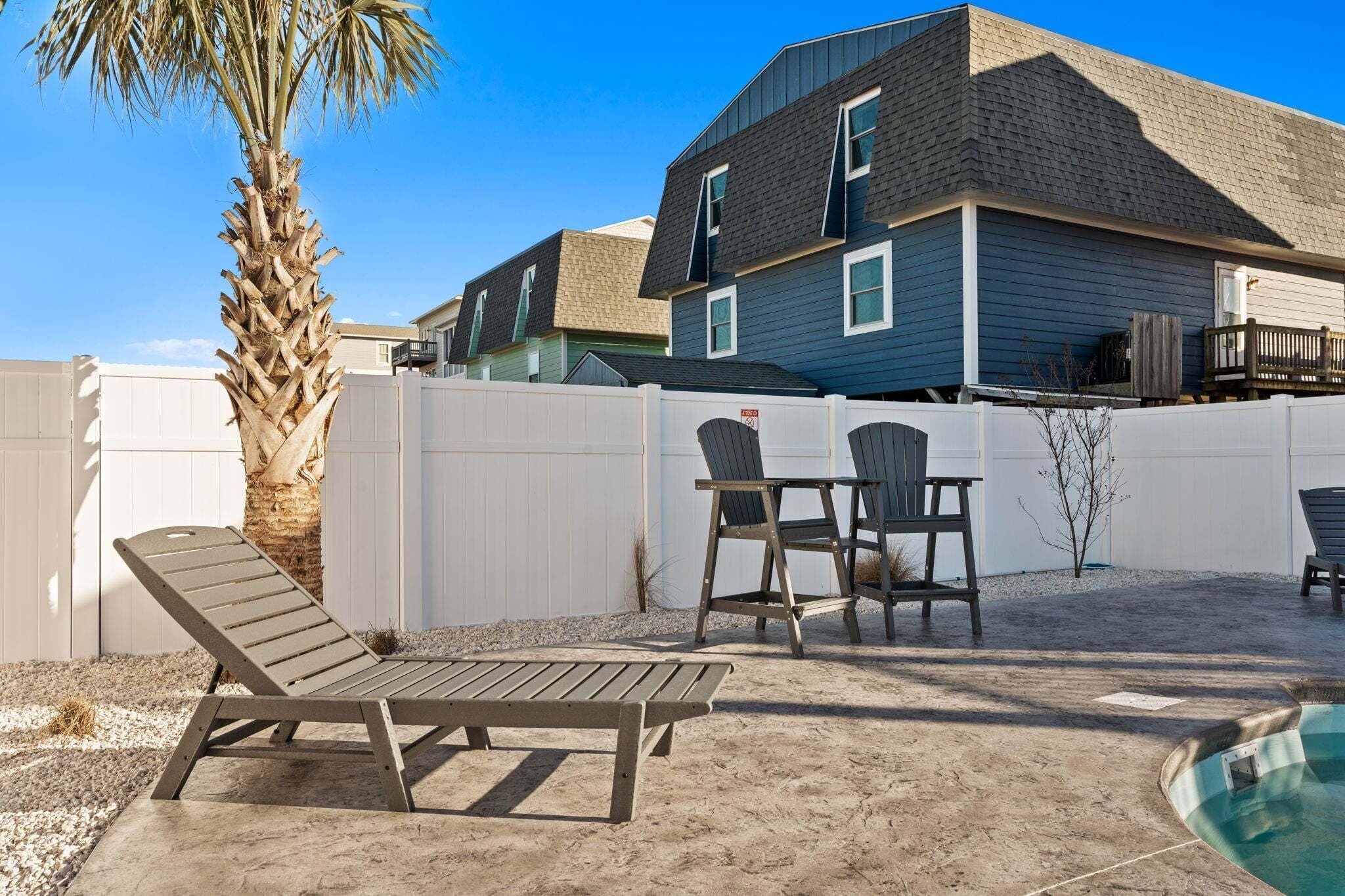 A backyard with a pool, a palm tree, and a house with a white fence.