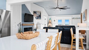Cozinha moderna com uma ilha grande, área de jantar com mesa branca e cadeiras de madeira, sofá preto e ventilador de teto.