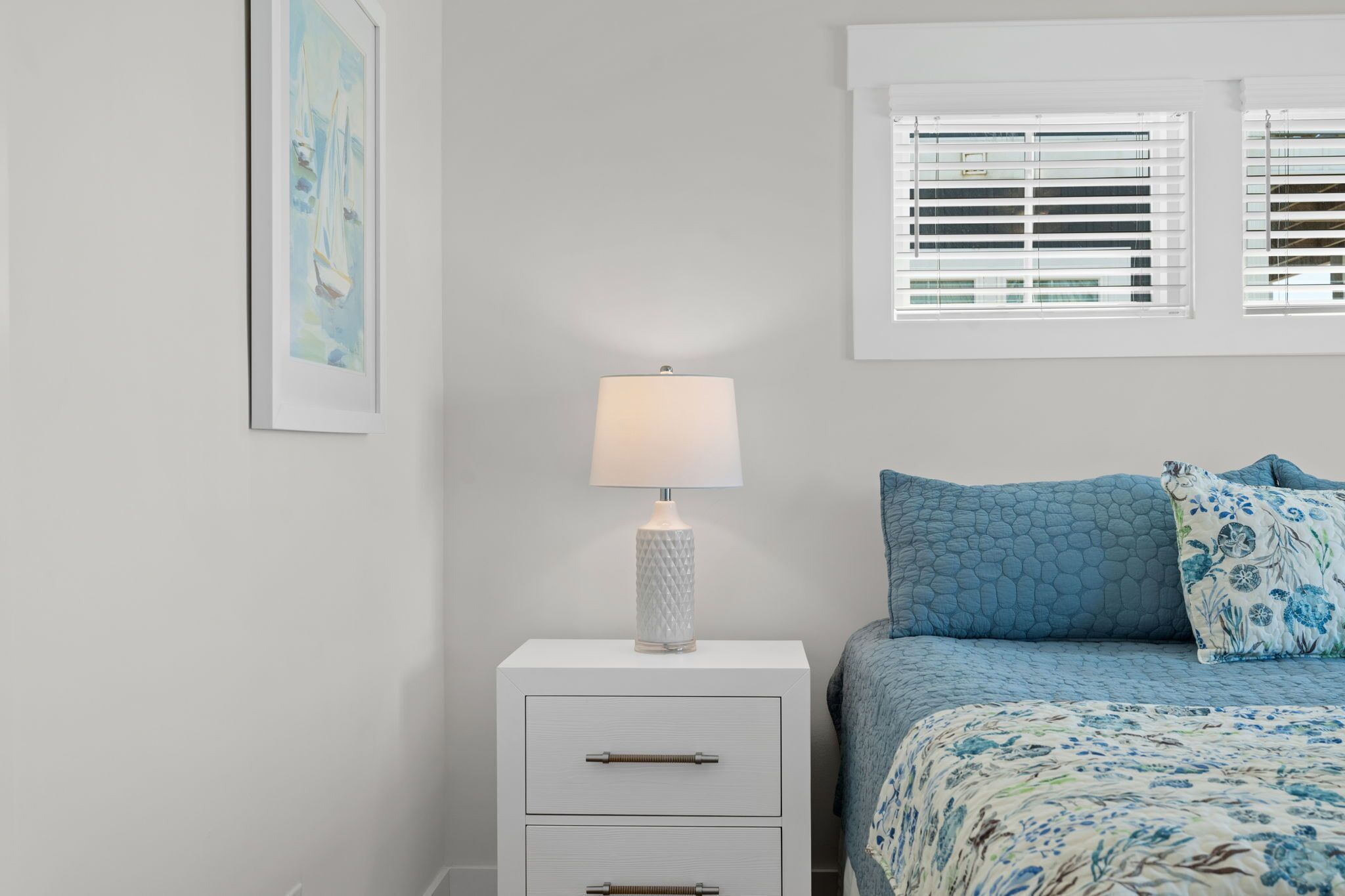 Un dormitorio con cama, una mesita de noche con lámpara, un cuadro enmarcado en la pared y una ventana con persianas.