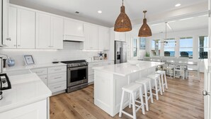 Uma cozinha moderna com armários brancos, uma ilha central com banquetas, eletrodomésticos de aço inoxidável e uma grande janela com vista para o oceano.