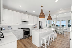 A modern kitchen with white cabinetry, a central island with bar stools, stainless steel appliances, and a large window overlooking the ocean.