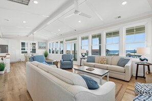 A spacious living area with a large sofa, armchairs, and a coffee table. There are multiple windows providing a view of the outside.