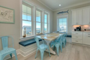 A kitchen with a long table, white cabinets, and a view through large windows.