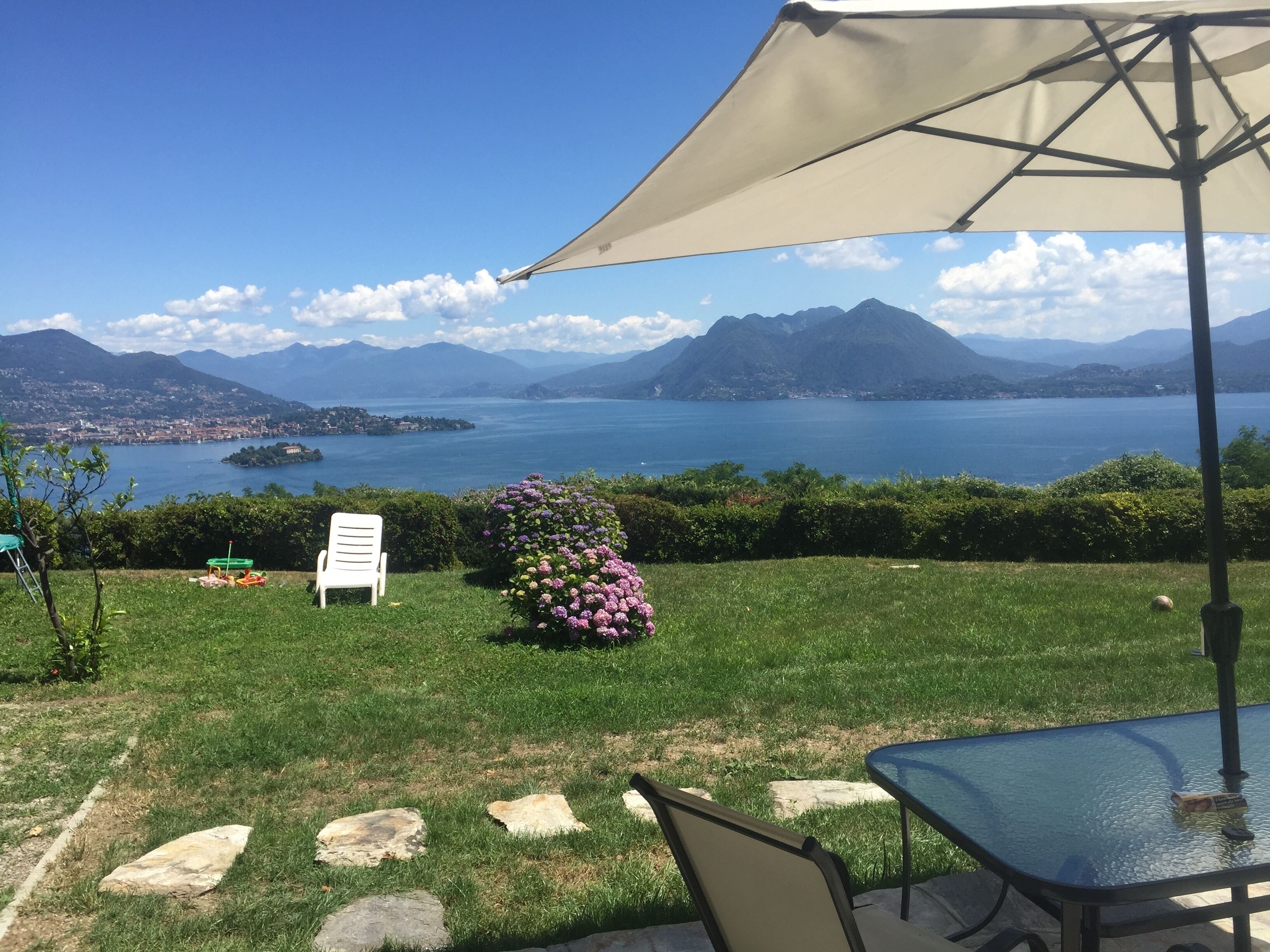 Un patio con tavolo e sedie, una sedia bianca, una pianta in vaso e vista su un lago con montagne sullo sfondo.
