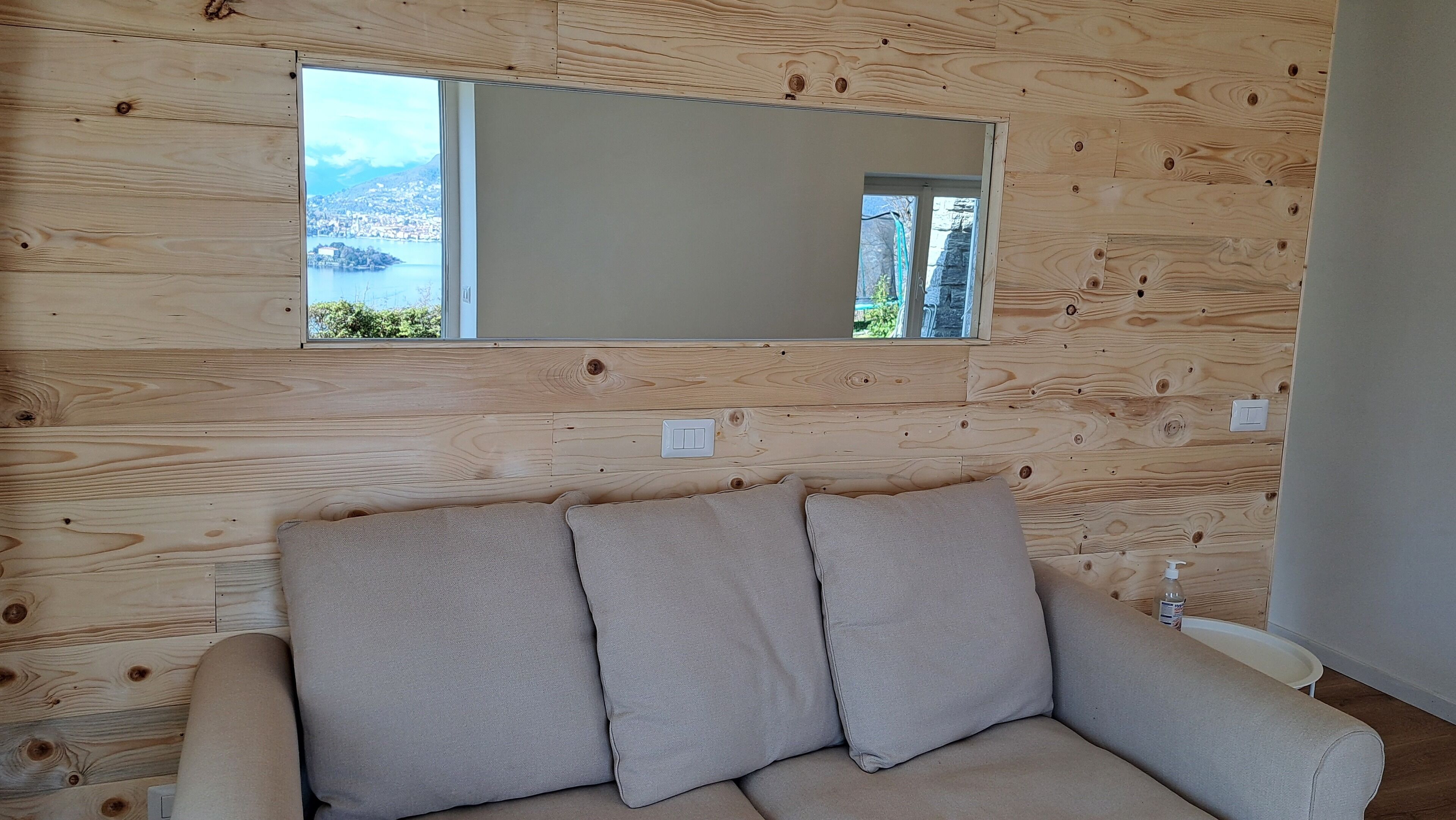 Una stanza con pannelli in legno sulla parete, un divano beige con cuscini, uno specchio grande e vista su un lago e degli edifici.