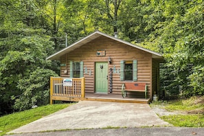 Une petite cabane en bois avec une porte verte, une pancarte indiquant « Bienvenue » et un banc à l’extérieur.