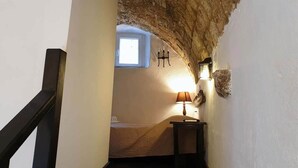 Un estrecho pasillo con techo de piedra rústica, una ventana pequeña y una cama con una lámpara.