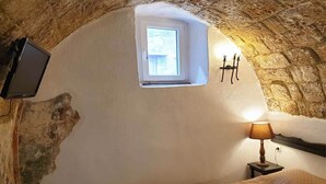 Un cuarto acogedor con pared de piedra, una ventana pequeña, cama con manta blanca, lámpara de noche y televisor de pantalla plana montado en la pared.