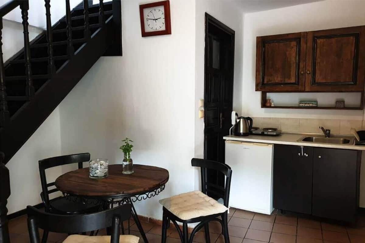 A dining area with a round table, two chairs, a clock on the wall, and a kitchen with cabinets and a stove.
