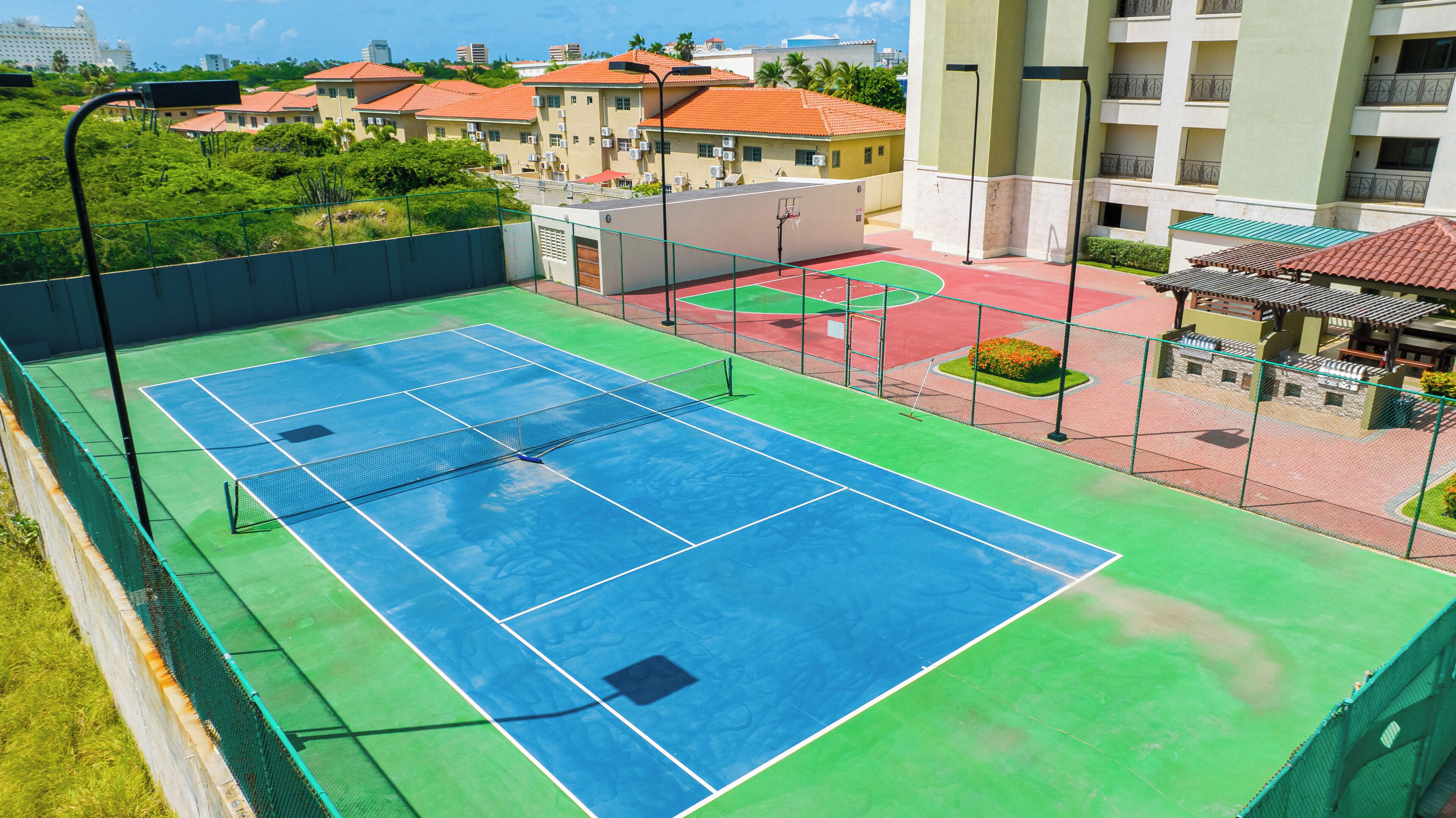 En tennisbane med net, omgivet af et grønt hegn, og tilstødende bygninger med røde tagsten.
