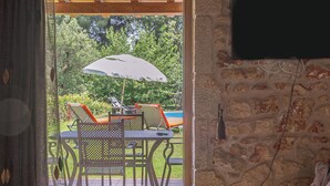 Une pièce chaleureuse avec un mur en pierre, un plafond en bois, une table en verre, un canapé et une vue sur une terrasse extérieure agrémentée de chaises et d’un parasol.