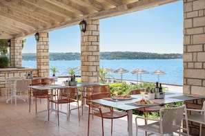 An outdoor dining area with stone pillars, wicker chairs, and tables overlooking a body of water. - Aminess Planet Camping Holiday Homes Sirena (Novigrad)