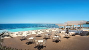 A rooftop bar with outdoor seating overlooking a pool and the sea.