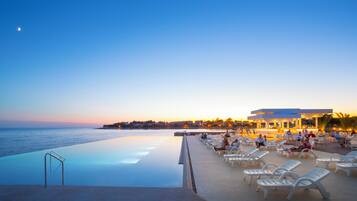 An infinity pool with a view of the sea, lounge chairs, and a building with a bar.