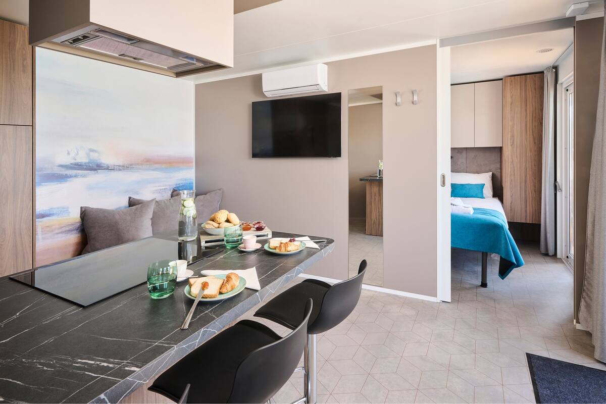 Una habitación de hotel moderna con zona de comedor, cama con cubrecama azul, televisor de pantalla plana y un cuadro de un paisaje.