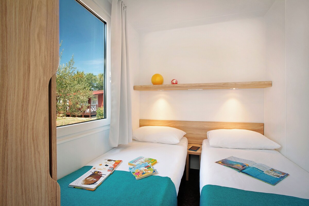 A hotel room with two single beds, a wooden headboard, a small shelf with decorative items, and a window with a view of greenery and a building outside.