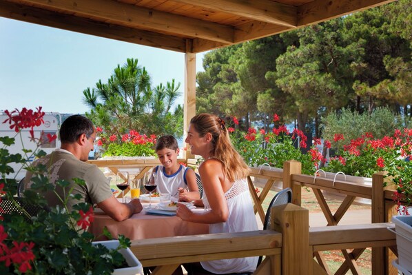 A wooden outdoor dining area with a covered roof, surrounded by lush greenery and red flowers. - Aminess Planet Camping Holiday Homes Sirena (Novigrad)