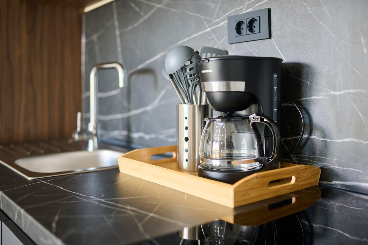 A kitchen with a coffee maker, a glass carafe, and utensils on a wooden tray.