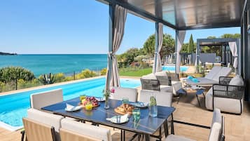 Un área de comedor junto a la piscina con vista al mar, mobiliario exterior y una mesa servida con comida y bebidas.