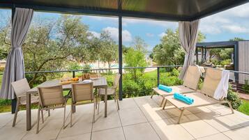A covered outdoor area with a table and chairs, a lounge area with sun loungers, and a view of a pool and greenery.