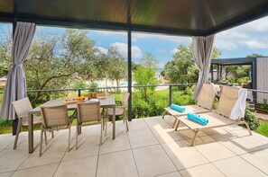 A covered outdoor area with a table and chairs, a lounge area with sun loungers, and a view of a pool and greenery. - Aminess Planet Camping Holiday Homes Sirena (Novigrad)