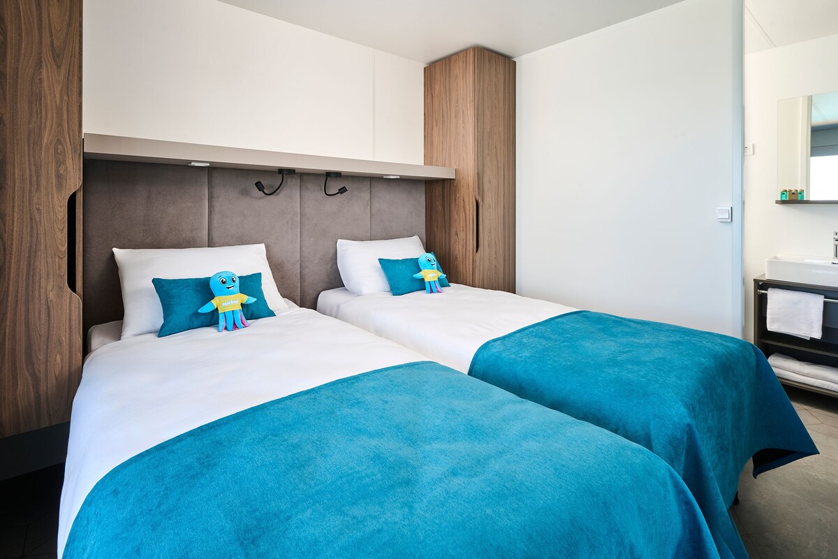 A hotel room with two beds, a wooden wardrobe, a small bathroom with a mirror and sink, and a blue plush toy on one of the beds.