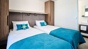 Habitación de hotel con dos camas, un armario de madera, un baño pequeño con espejo y lavamanos, y un peluche azul sobre una de las camas.