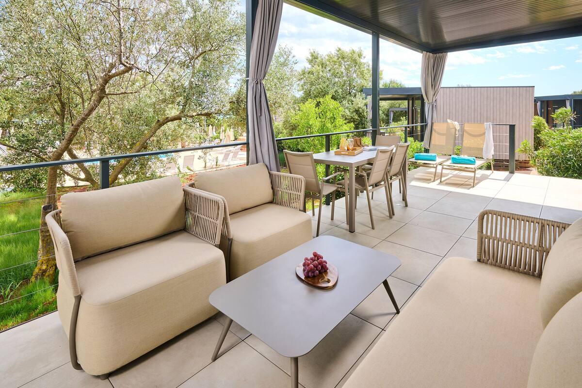 Un espacio al aire libre cubierto con sofás, una mesa y sillas, con vista a un jardín y un edificio.