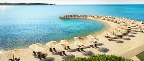 A beach with sun umbrellas and lounge chairs arranged in rows, a clear blue sea, and a rocky outcrop in the distance.