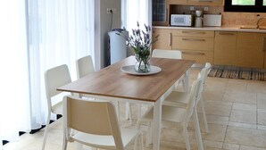 Een moderne eethoek met een houten tafel en witte stoelen, een vaas met bloemen en een keuken op de achtergrond.