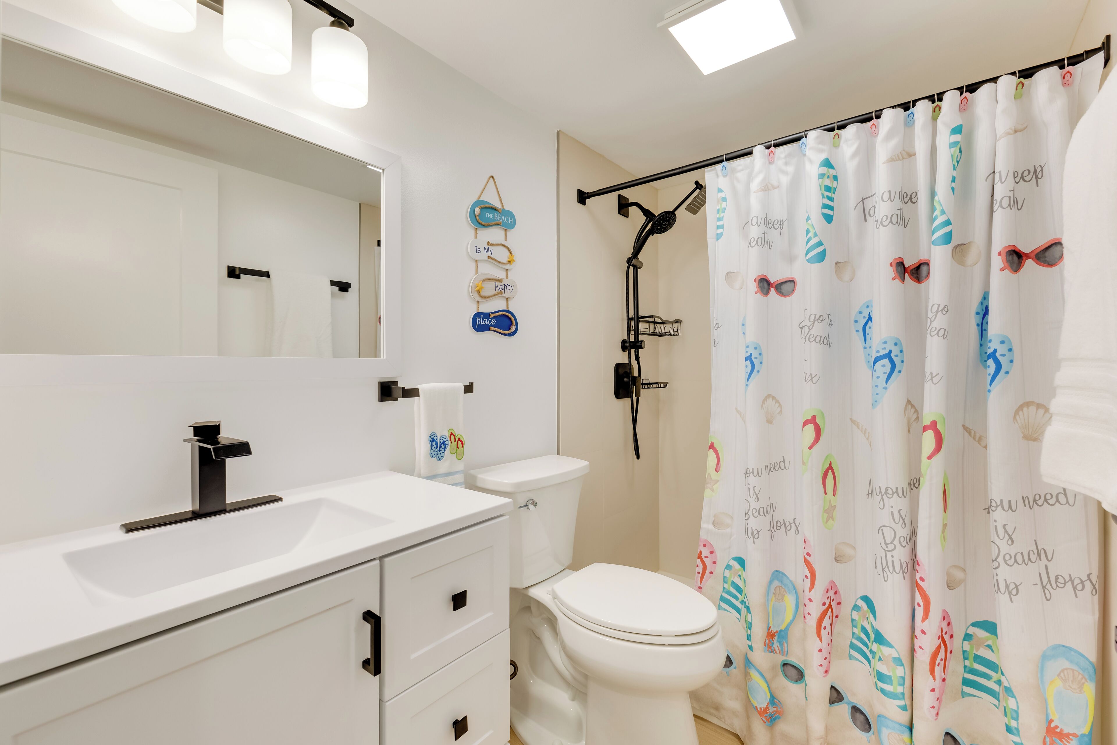 Ein Badezimmer mit weißem Waschbecken, Toilette, Dusche inklusive schwarzem Duschschlauch und Deckenleuchte.