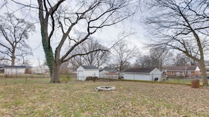 A fenced yard with leaf-covered ground, a tree with ivy, and a white building in the background.